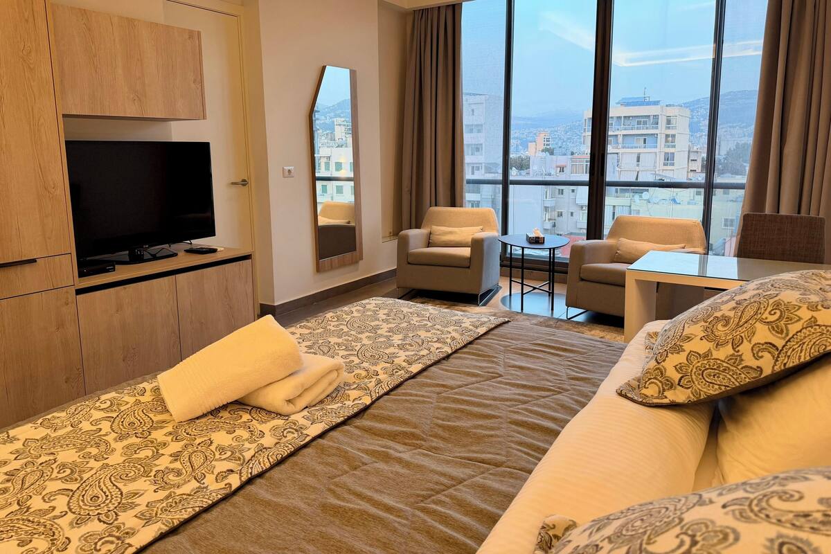 A hotel room with a large bed, two armchairs, a small table, a TV, and a view of the city through large windows.