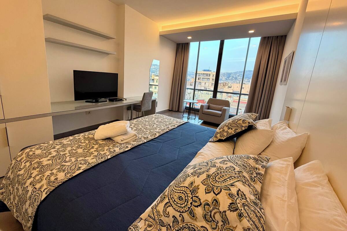 A hotel room with two beds, a desk with a TV, a chair, and a view of the city through large windows.