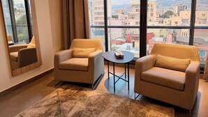 A modern hotel room with two beige armchairs, a small round table, and a large window offering a city view. - Unit for Living (Beirut)