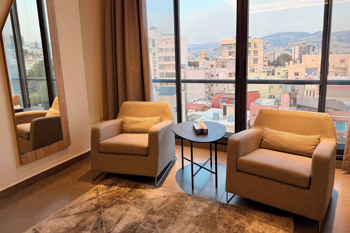 A modern hotel room with two beige armchairs, a small round table, and a large window offering a city view.