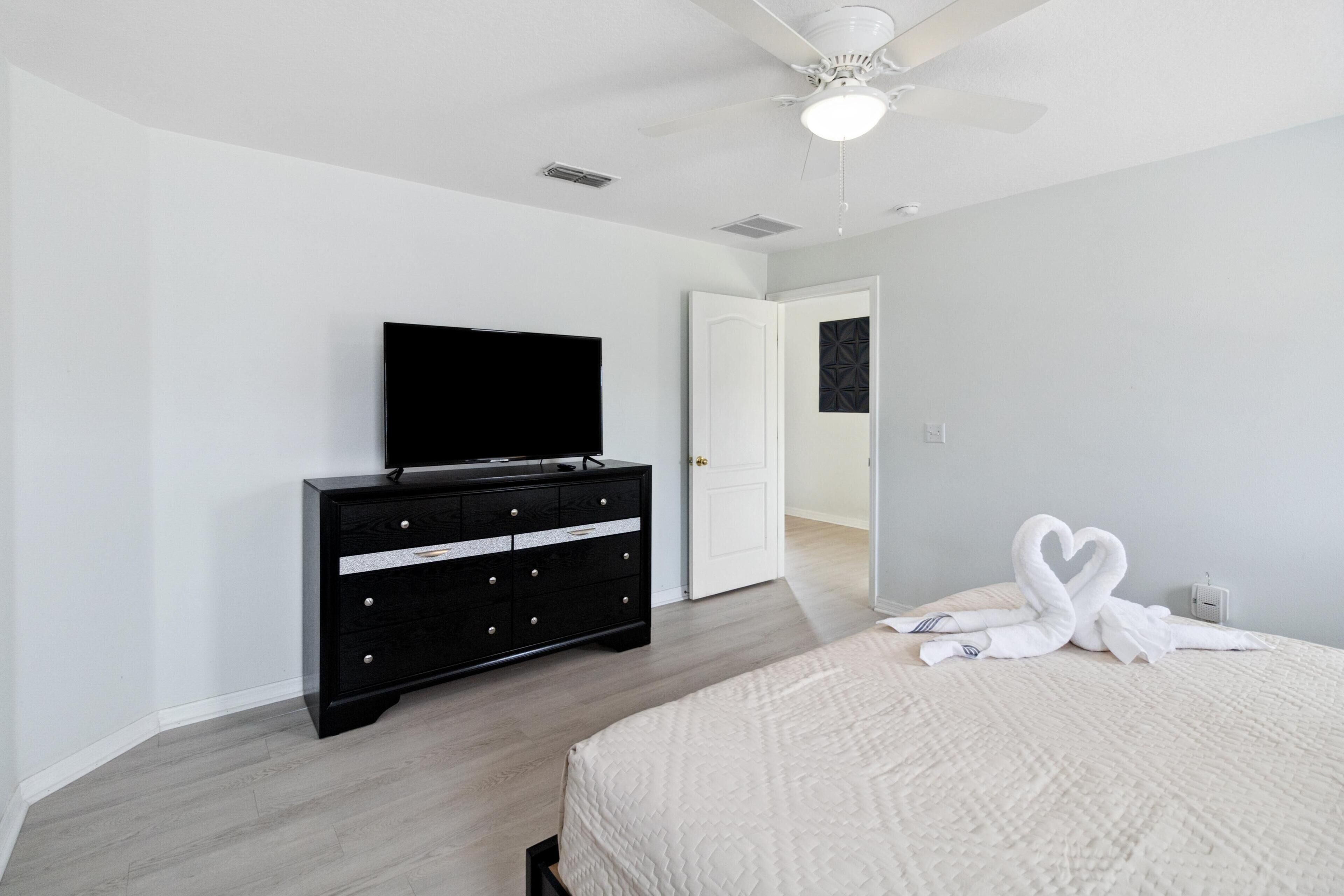 A bedroom with a bed, a dresser, a flat-screen TV, a ceiling fan, and a door leading to another room.