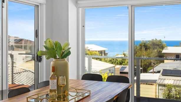 A dining area with a table set for two, a bottle of wine, and a glass on the table. A vase with green leaves is on the table. The room has large windows with a view of the sea and buildings.