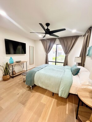 Un dormitorio moderno con cama, ventilador de techo, televisor, mesita de noche, lámpara y ventana con cortinas.