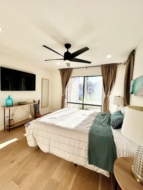 Un dormitorio con cama, ventilador de techo, televisión y ventana con cortinas.