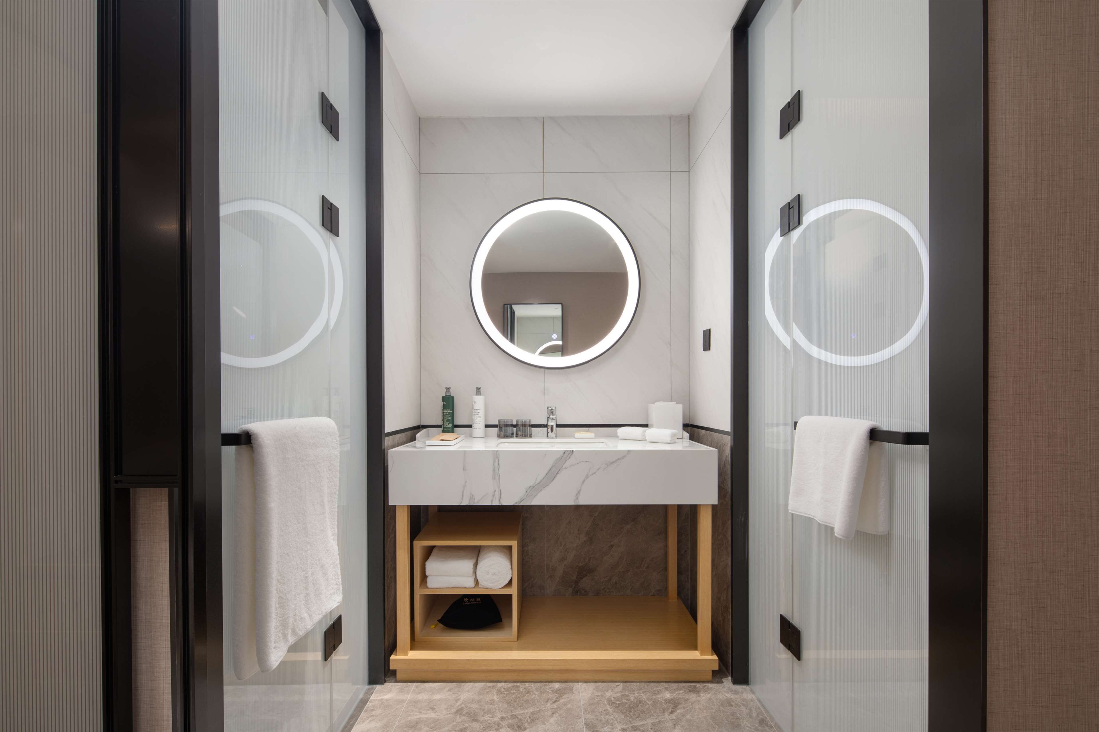 Une salle de bain moderne avec un meuble-lavabo en marbre, deux miroirs ronds et une étagère en bois où sont posées des serviettes.