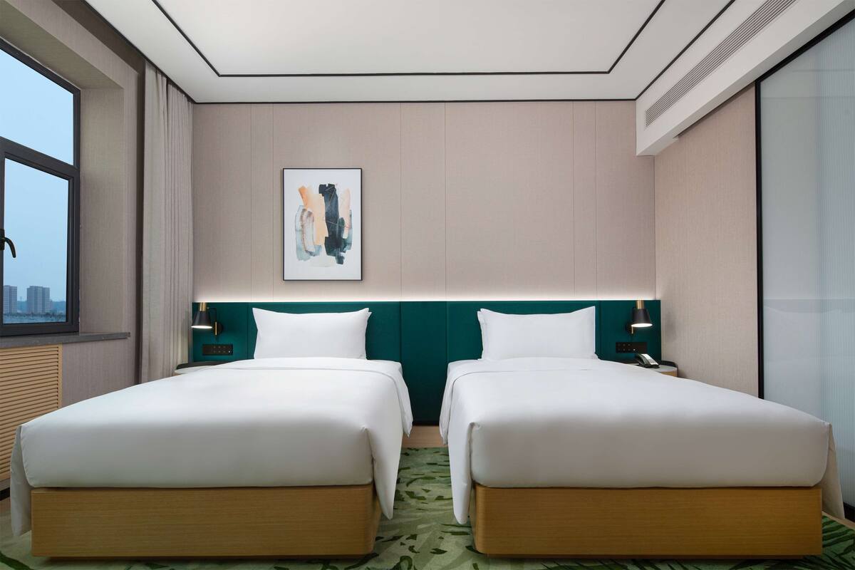 Ein Hotelzimmer mit zwei Betten, Blick auf die Stadt, einem modernen Kopfteil und einem gerahmten Kunstwerk an der Wand.