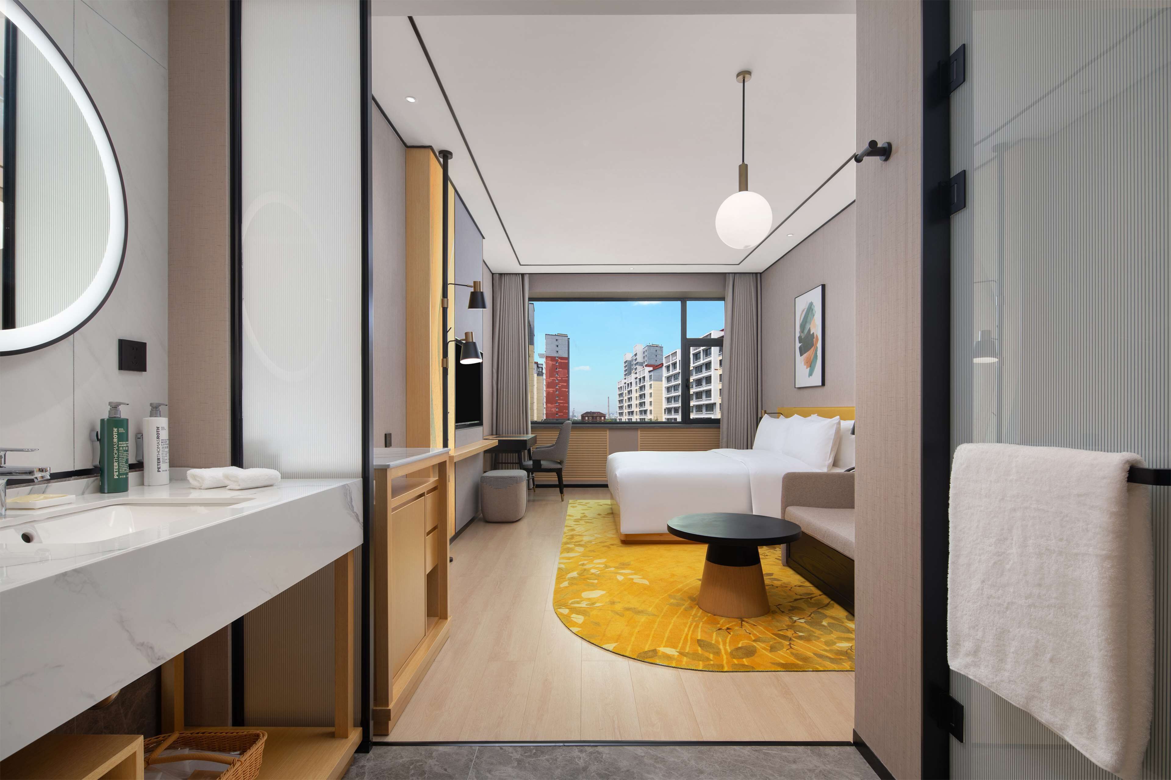 Una habitación de hotel moderna con un ventanal grande, una cama, un sofá, una mesa redonda y un baño con lavabo y espejo.