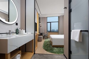 A hotel room with a marble bathroom, a bed with a green leaf-patterned rug, a city view, and a large mirror.