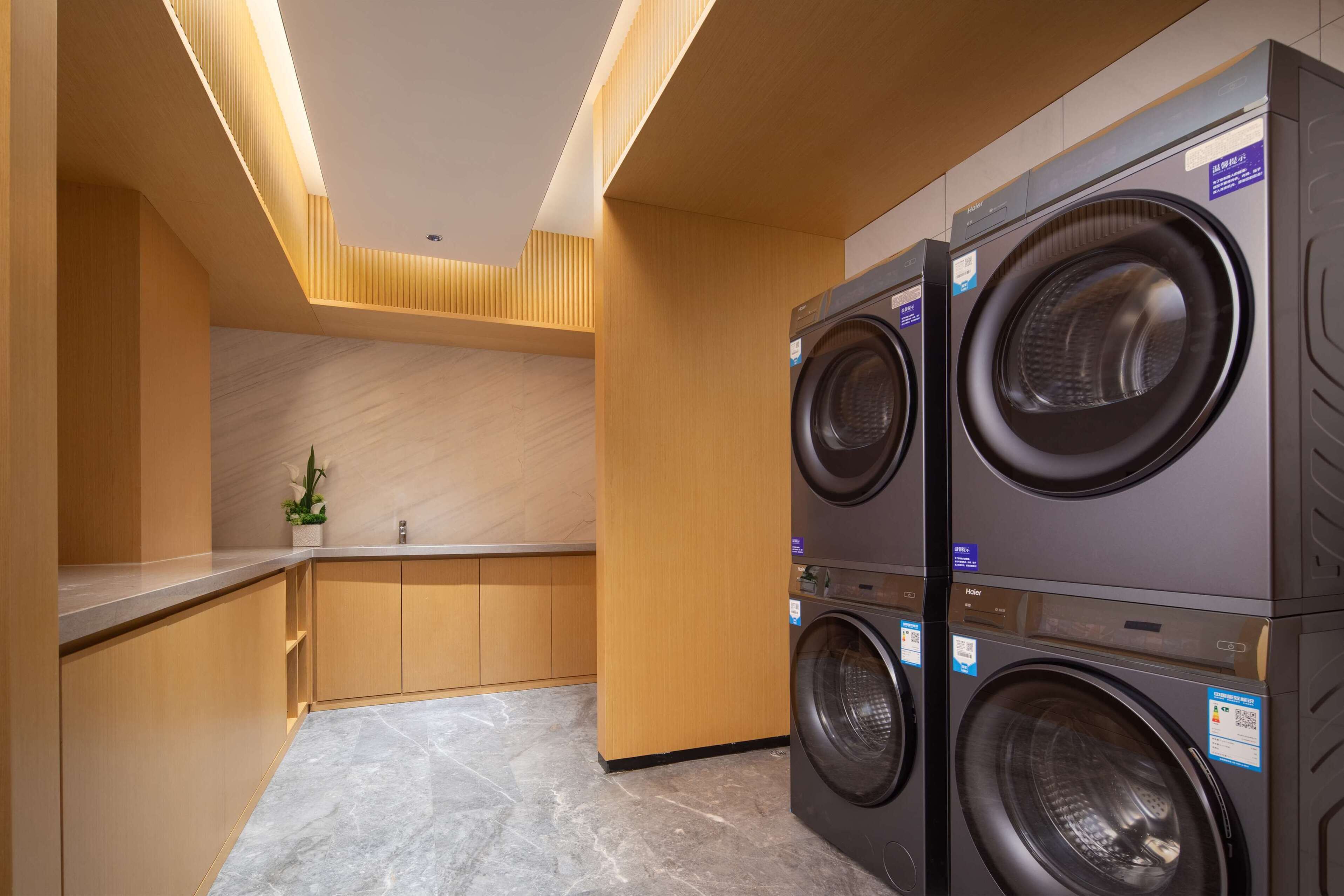 A modern laundry room with two stacked front-loading washing machines, wooden cabinetry, and a marble-tiled floor.
