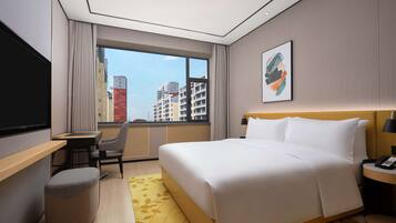 Habitación de hotel con cama, escritorio, silla, televisor y ventana con vistas a edificios.