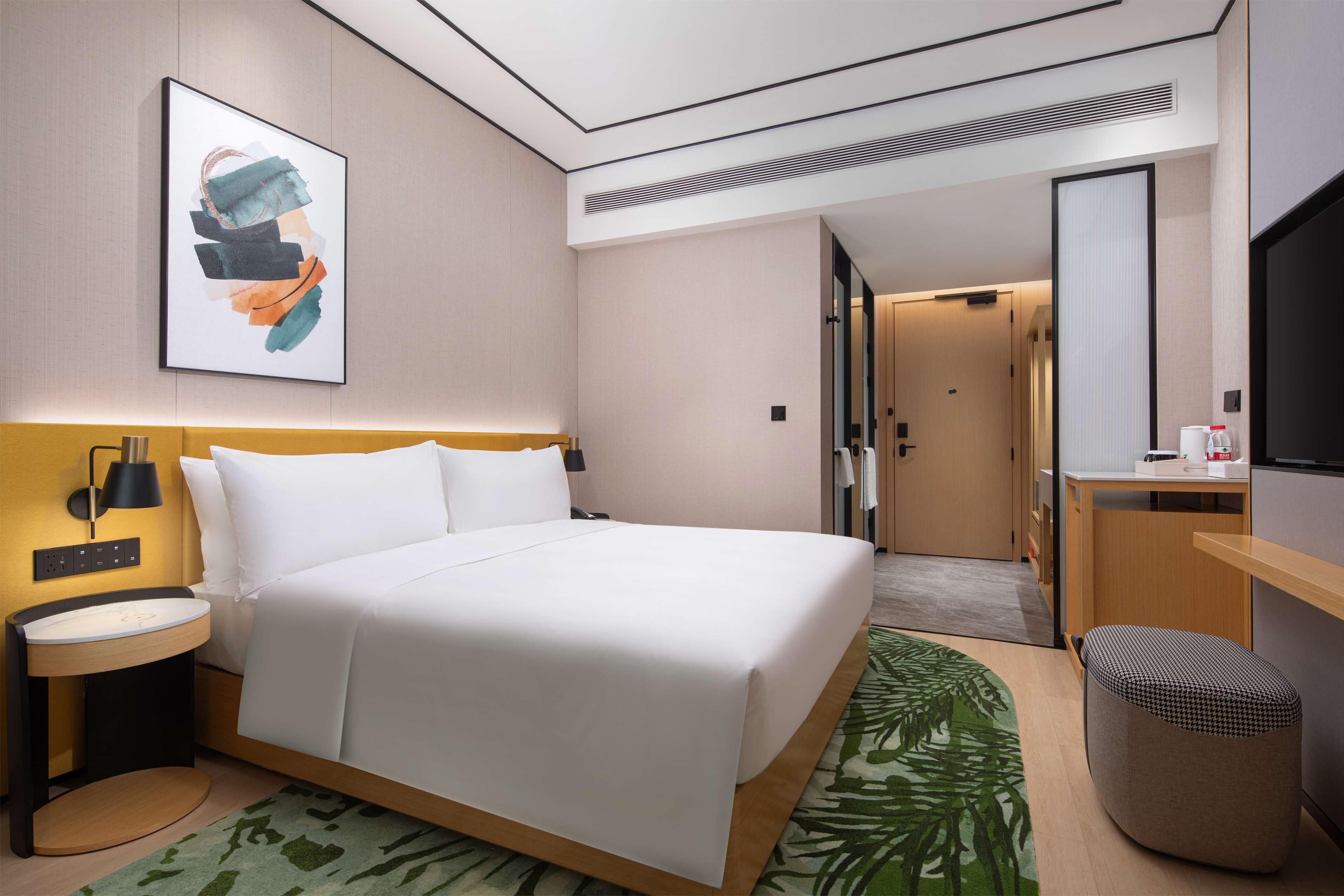 A modern hotel room with a large bed, bedside table, and a small desk with a chair.