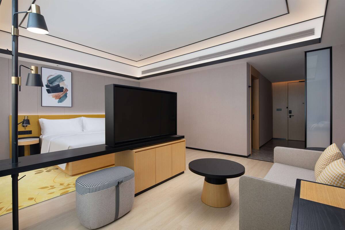 Ein modernes Hotelzimmer mit einem Bett, einem Flachbildfernseher, einer Couch, einem kleinen runden Tisch und einer Bank.