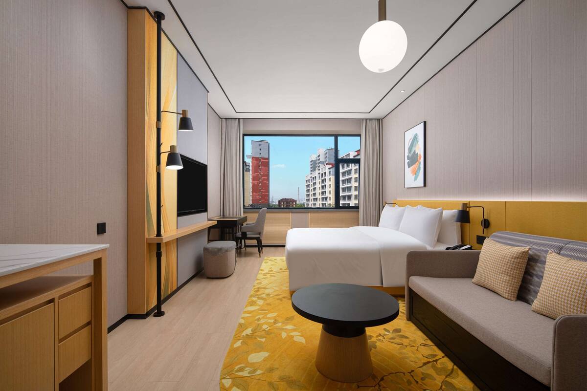 Ein Hotelzimmer mit einem Bett, einem Sofa, einem Schreibtisch, einem Fernseher und einem großen Fenster mit Blick auf die Stadt.