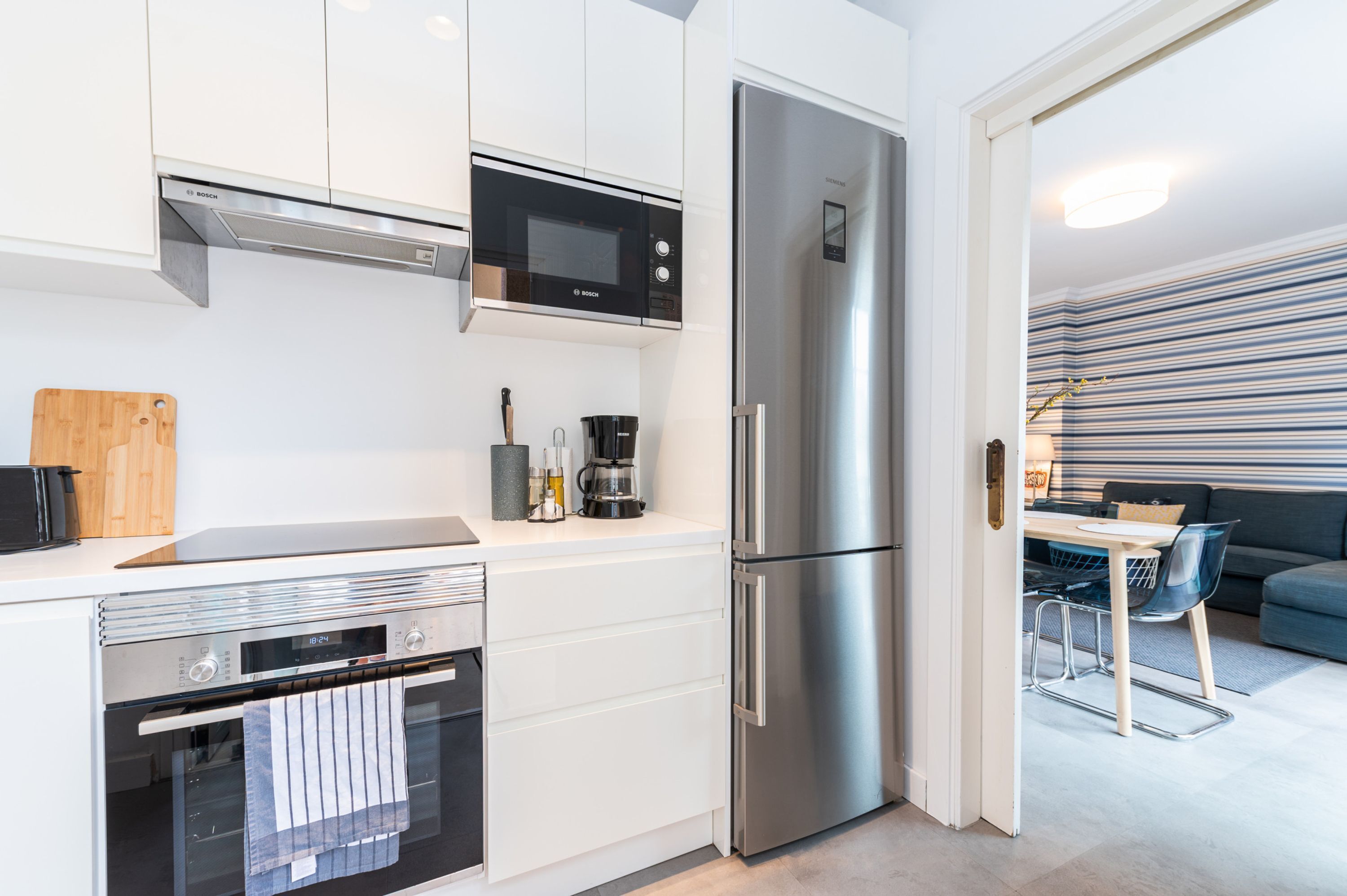 A modern kitchen with white cabinets, a Bosch microwave, a stainless steel refrigerator, and a dining area with a striped wall and blue sofa.