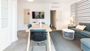 En moderne stue med spiseplads, et fladskærms-tv på væggen og en grå sofa med et mønstret pude.
