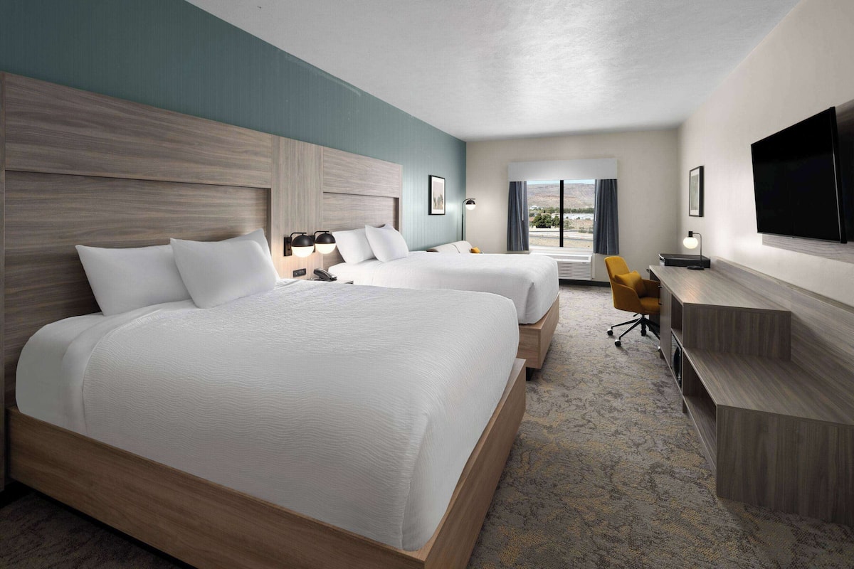 Quarto de hotel com duas camas, uma escrivaninha, uma televisão e vista para o exterior.