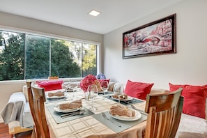 Un coin repas avec une table dressée pour quatre personnes, une fenêtre donnant sur les arbres et un tableau accroché au mur.