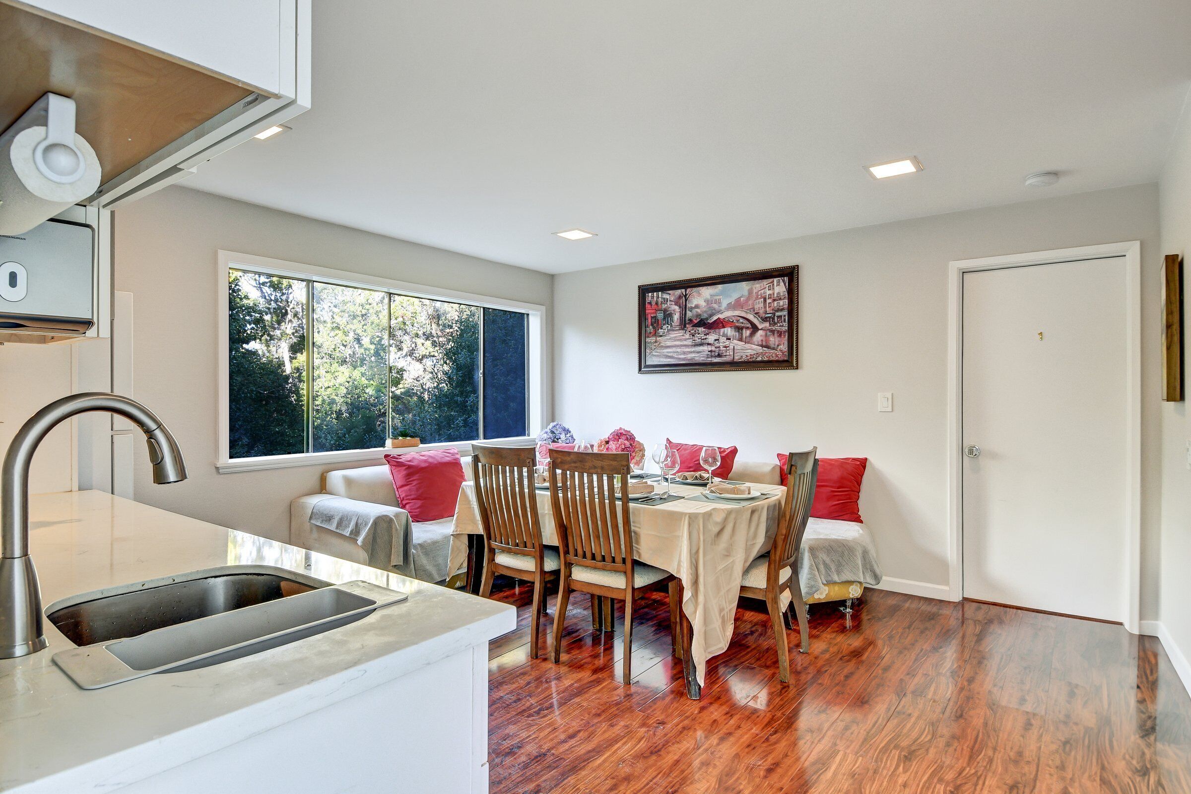 A dining area with a table set for four, wooden chairs, a window with a view of trees, and a painting on the wall.