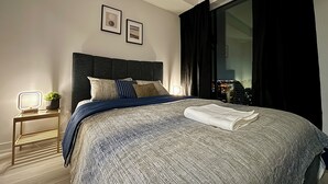 A modern bedroom with a large bed, a bedside table with a lamp, and a view of the city at night.