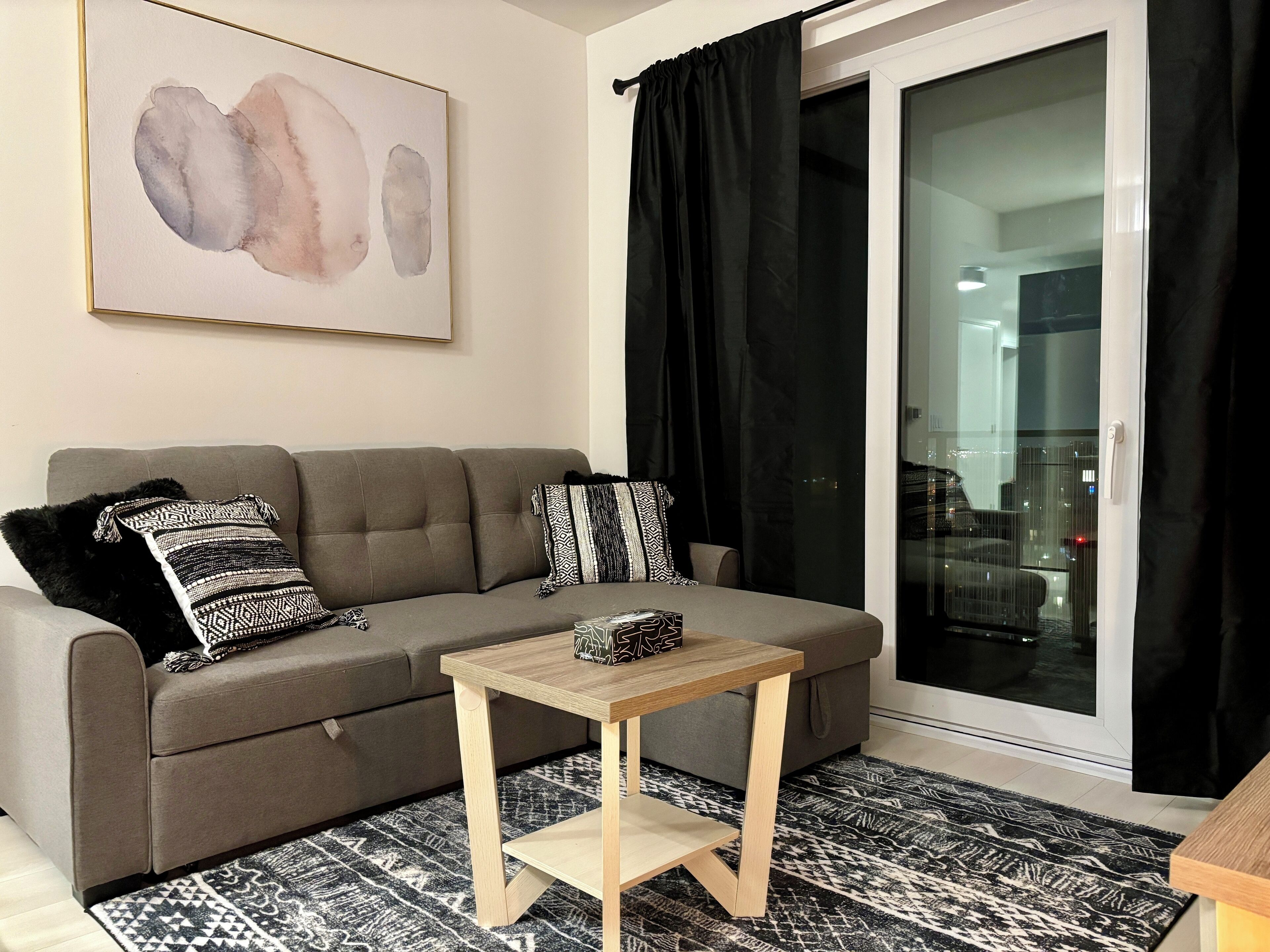 A living room with a grey sofa, patterned cushions, a small wooden table, and a black and white patterned rug.