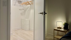 A room with a bed, a bedside table with a lamp, a small plant, and a wall-mounted shelf with folded towels.