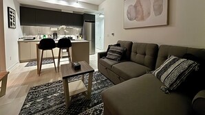 A modern living room with a sofa, a coffee table, and a patterned rug. There is a kitchen area with a refrigerator and a dining table with chairs.