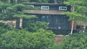 Une cabane moderne dotée d’une grande baie vitrée, entourée d’une forêt dense et bordée par un plan d’eau.