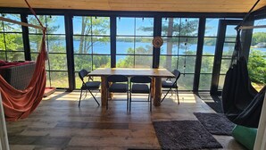 Une table à manger en bois, accompagnée de quatre chaises, un hamac et une vue sur un lac, visible à travers de grandes fenêtres.