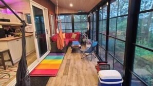 L’intérieur chaleureux d’un chalet, avec un plancher en bois, un tapis coloré, un hamac et une grande fenêtre donnant sur le paysage extérieur.