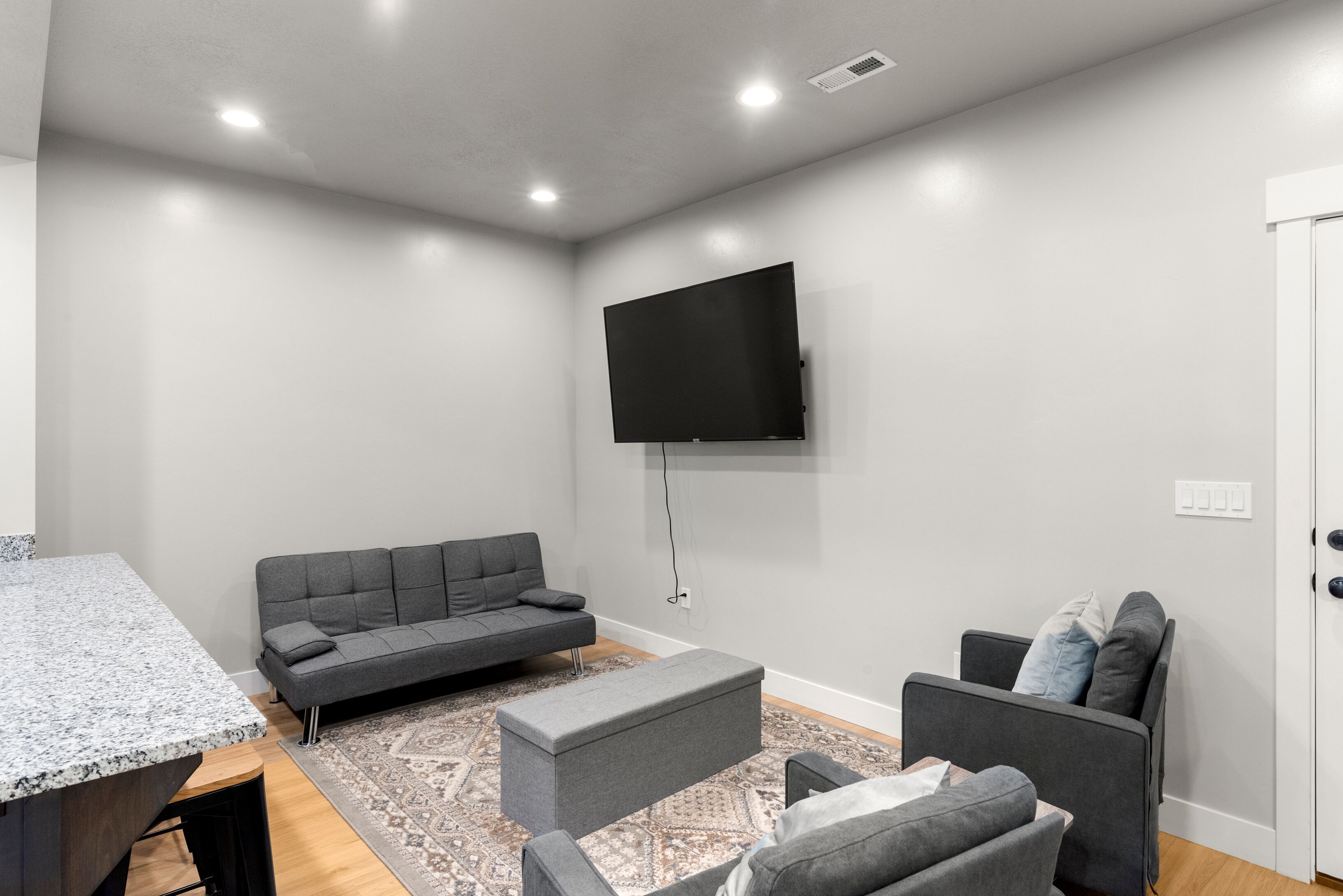 Uma sala de estar moderna com um sofá cinzento, uma poltrona cinzenta, um banquinho pequeno e uma televisão de tela plana fixada na parede.