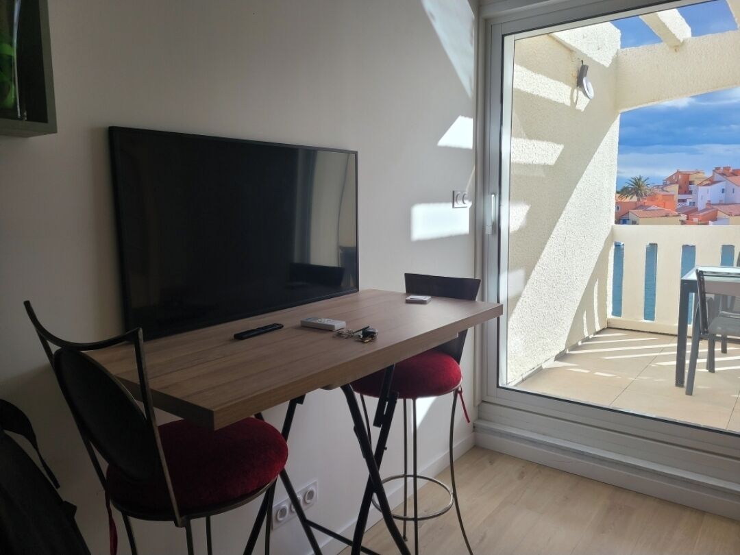 A room with a TV on a wooden table, red cushioned chairs, and a view of a building through a glass door.