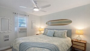 A bedroom with a bed, two bedside tables with lamps, a ceiling fan, a window with blinds, and a decorative mirror on the wall. - Walk to the Beach! Firepit, games, pets welcome, bring your boat! (Edisto Island)