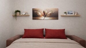 A bedroom with a bed, two red pillows, a beige blanket, a wall-mounted painting, and two shelves with decorative items.