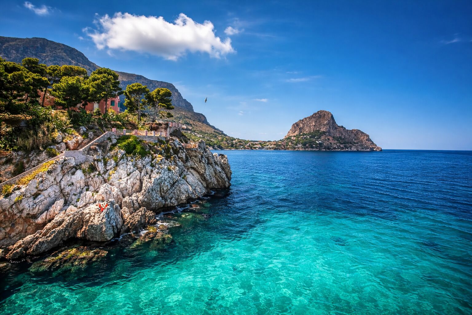 A coastal landscape with clear turquoise waters, rocky cliffs, and a prominent mountain in the distance.