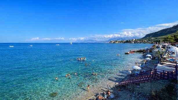 A coastal beach scene with clear water, people swimming and sunbathing, and a rocky shoreline with umbrellas and beach chairs.