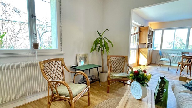 Ein geräumiges Wohnzimmer mit großem Fenster, Korbstühlen, einem Holztisch und einer Zimmerpflanze.