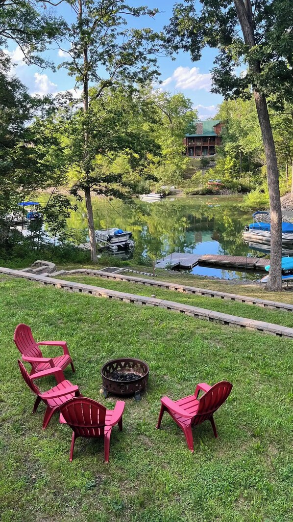A lakeside outdoor seating area with red chairs, a fire pit, and a wooden dock extending into the water.