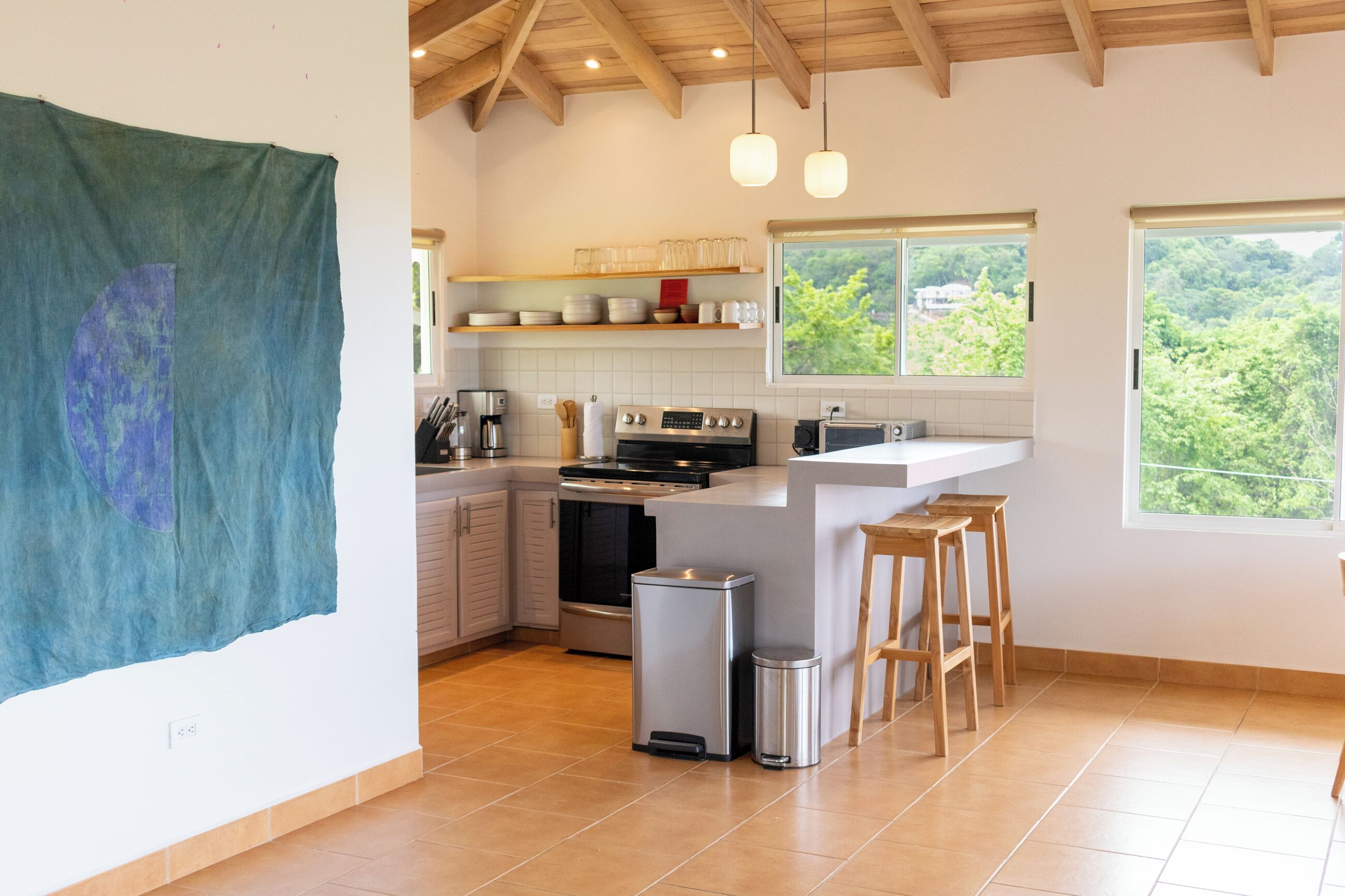 Een moderne keuken met een centraal eiland, houten krukken en uitzicht op groen door grote ramen.