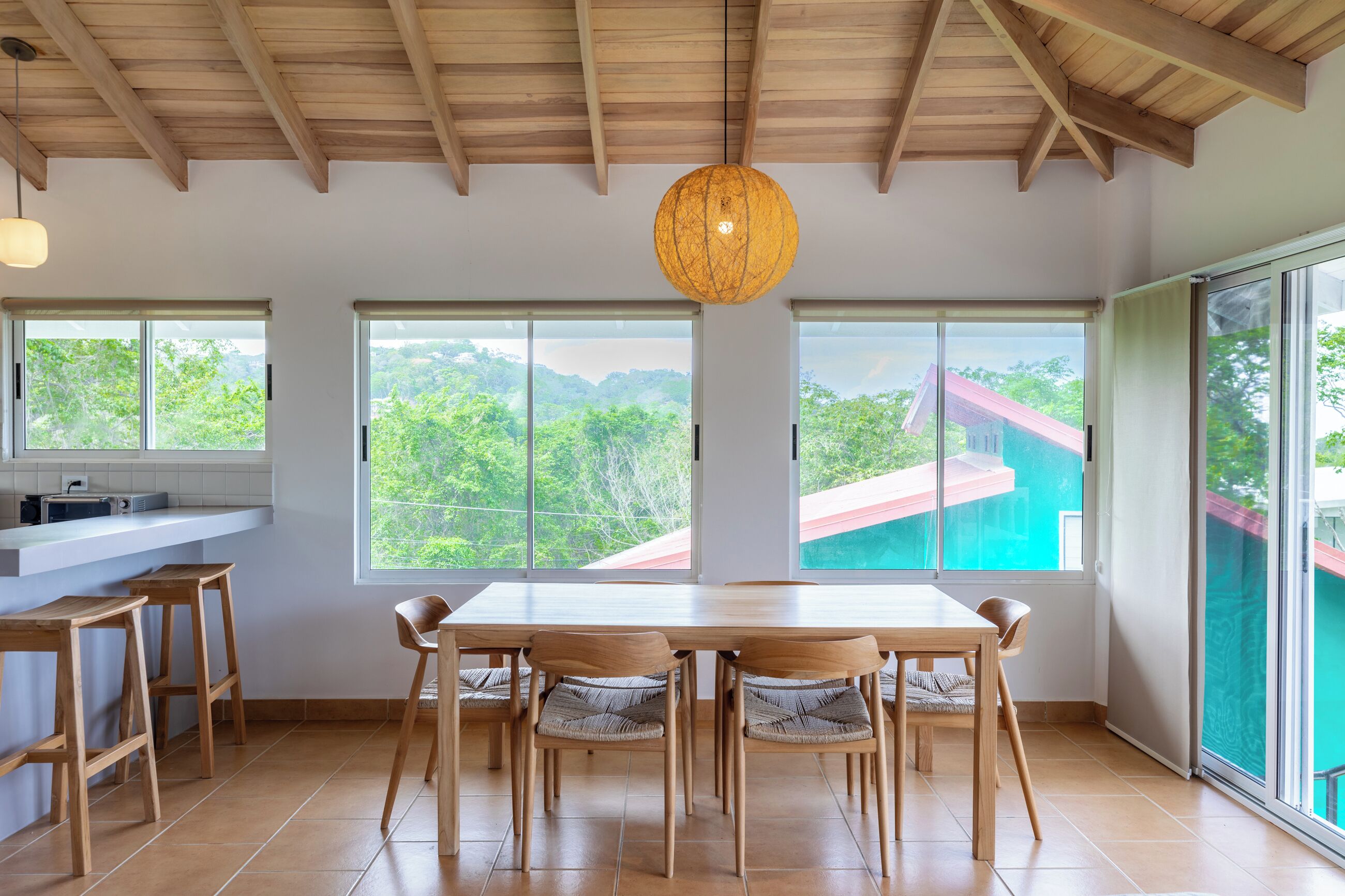 A spacious dining area with a wooden table and chairs, large windows offering a view of greenery and a building, and a hanging light fixture.