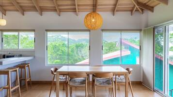 A spacious dining area with a wooden table and chairs, large windows offering a view of greenery and a building, and a hanging light fixture.