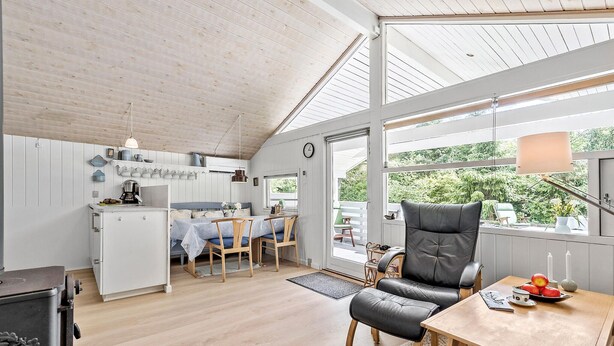 Um quarto acolhedor e bem iluminado, com teto de madeira, área de jantar com mesa e cadeiras, cozinha com fogão e grandes janelas com vista para o verde lá fora.