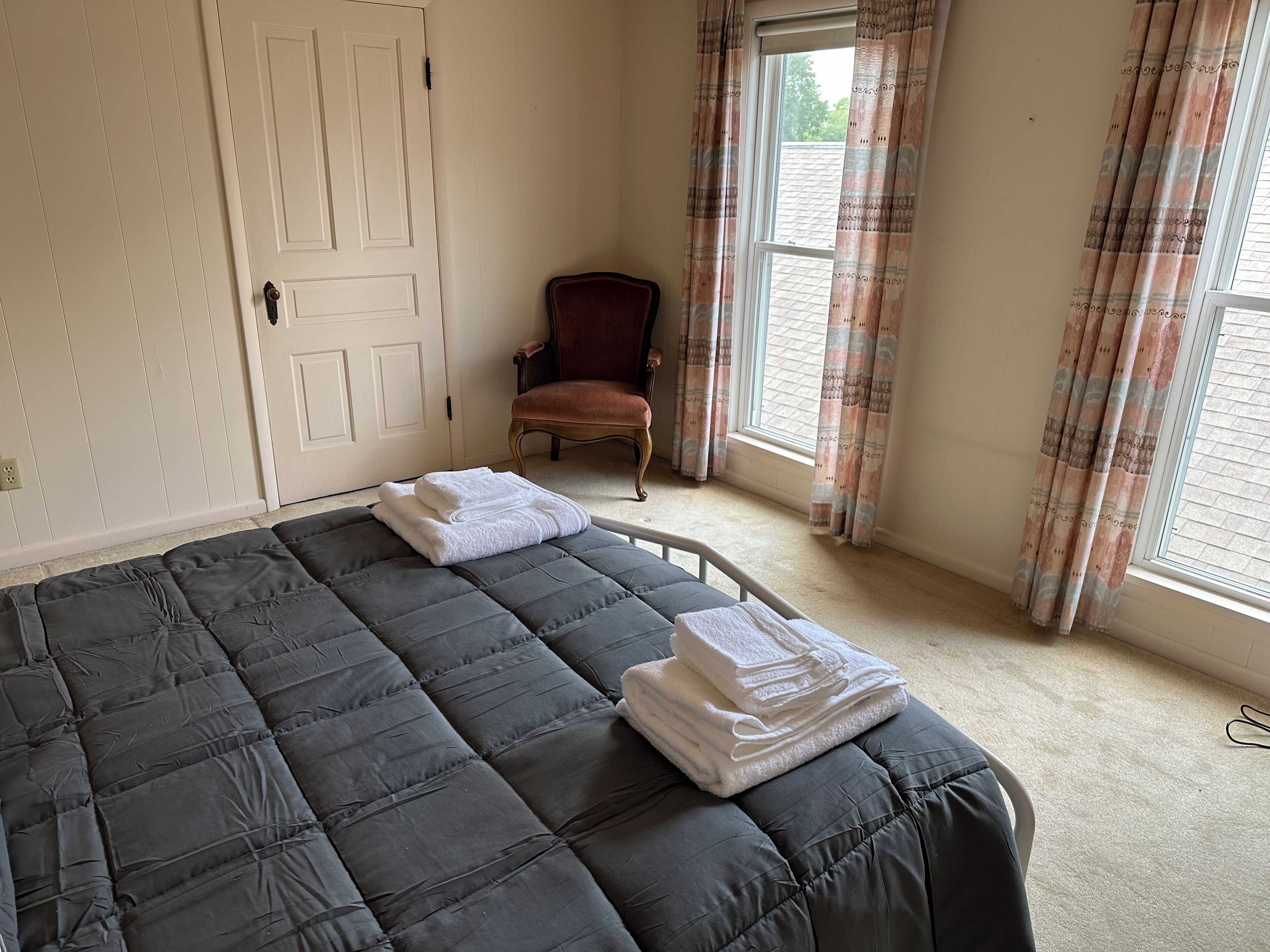 Un dormitorio con una cama grande, una silla y un ventanal con cortinas.