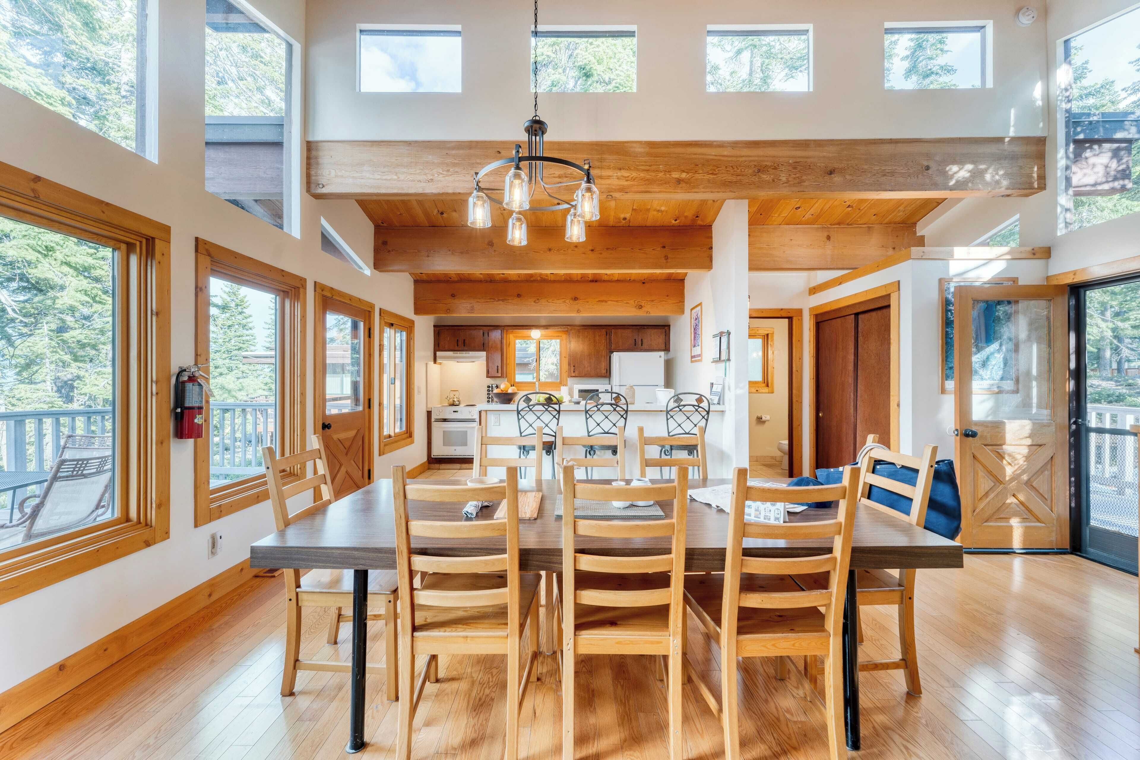 A spacious dining area with a wooden table and chairs, large windows, and a kitchen in the background.