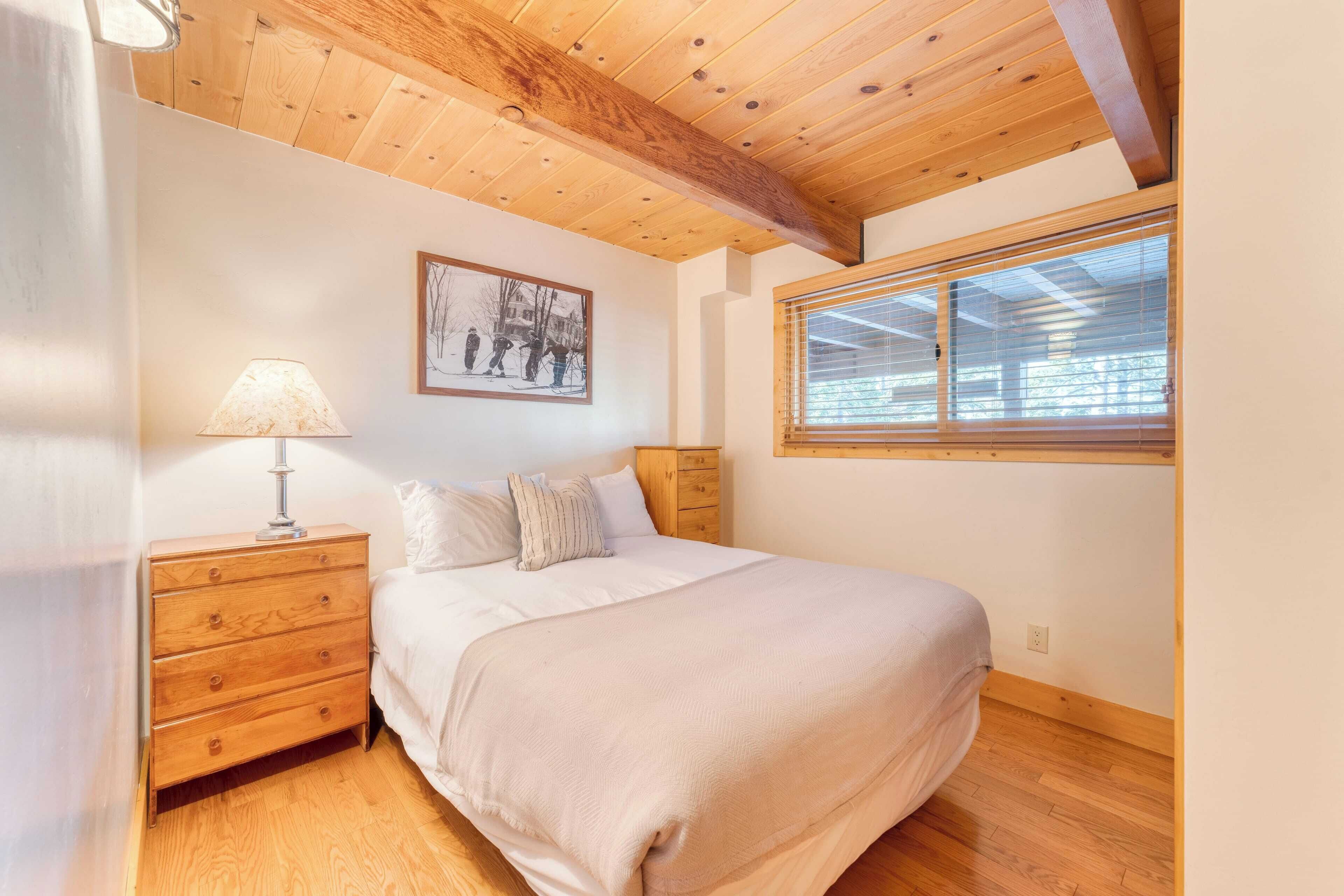 Een slaapkamer met een houten plafond, een bed met witte lakens, een nachtkastje met een lamp, een raam met jaloezieën, en een lijst met een afbeelding aan de muur.
