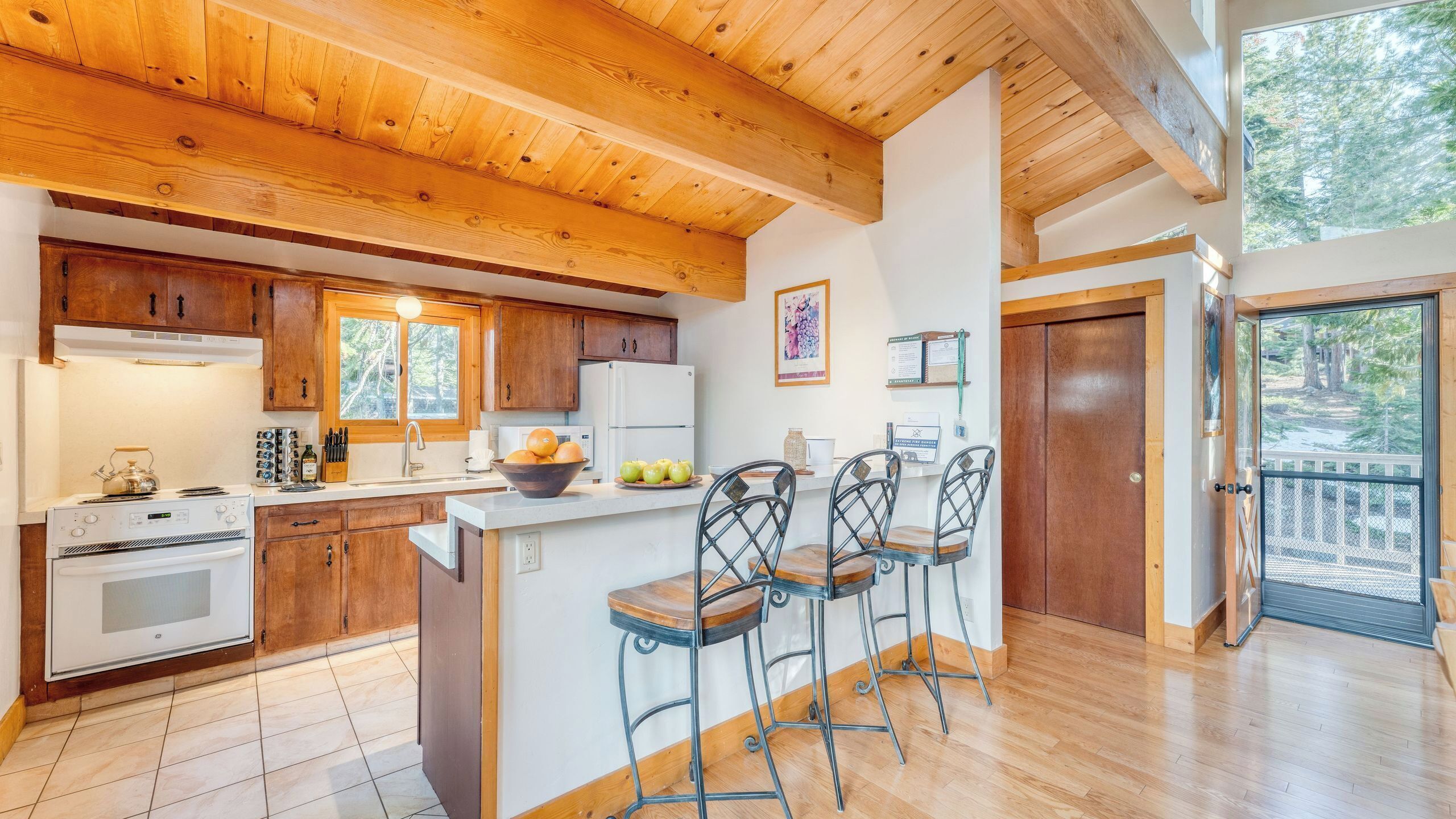 Cocina con gabinetes de madera, estufa y refrigerador. Hay dos taburetes de bar y una ventana con vista a árboles.