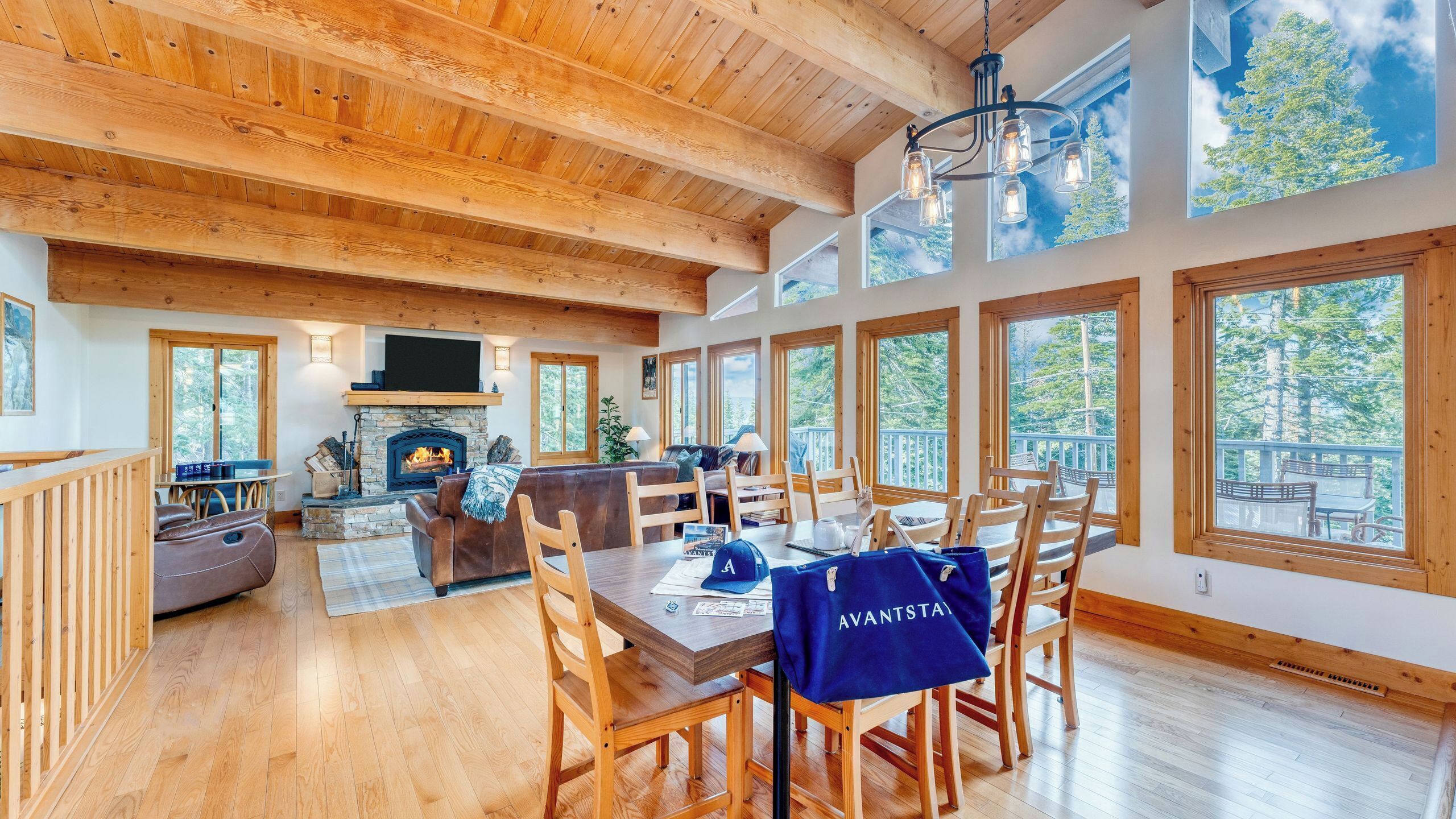 A spacious living area with a fireplace, wooden ceiling, and large windows offering views of trees.