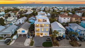 A coastal residential area with a prominent two-story house featuring a balcony, a driveway, and parked cars.