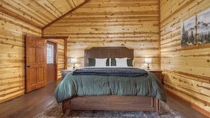 A log cabin bedroom with a wooden bed, two bedside lamps, a door, and a painting on the wall. - Evergreen Lodge by Avantstay Indoor Heated Pool + Game Room + Movie Theater + Hot Tub Mtn Views + Pigeon Forge (Sevierville)