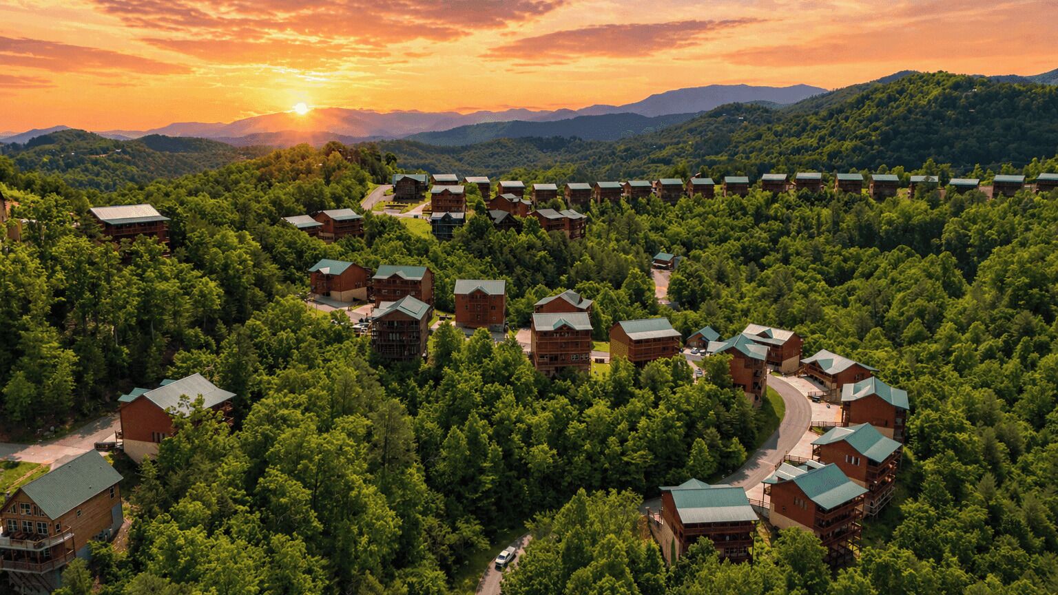 A resort with multiple lodge-style buildings surrounded by dense forest, connected by winding roads, and set against a mountainous backdrop.