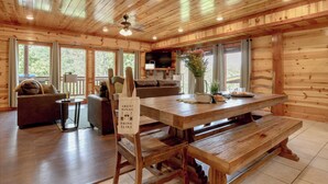 Interior de una cabaña de madera con una mesa de comedor, bancos, un sofá y un televisor.